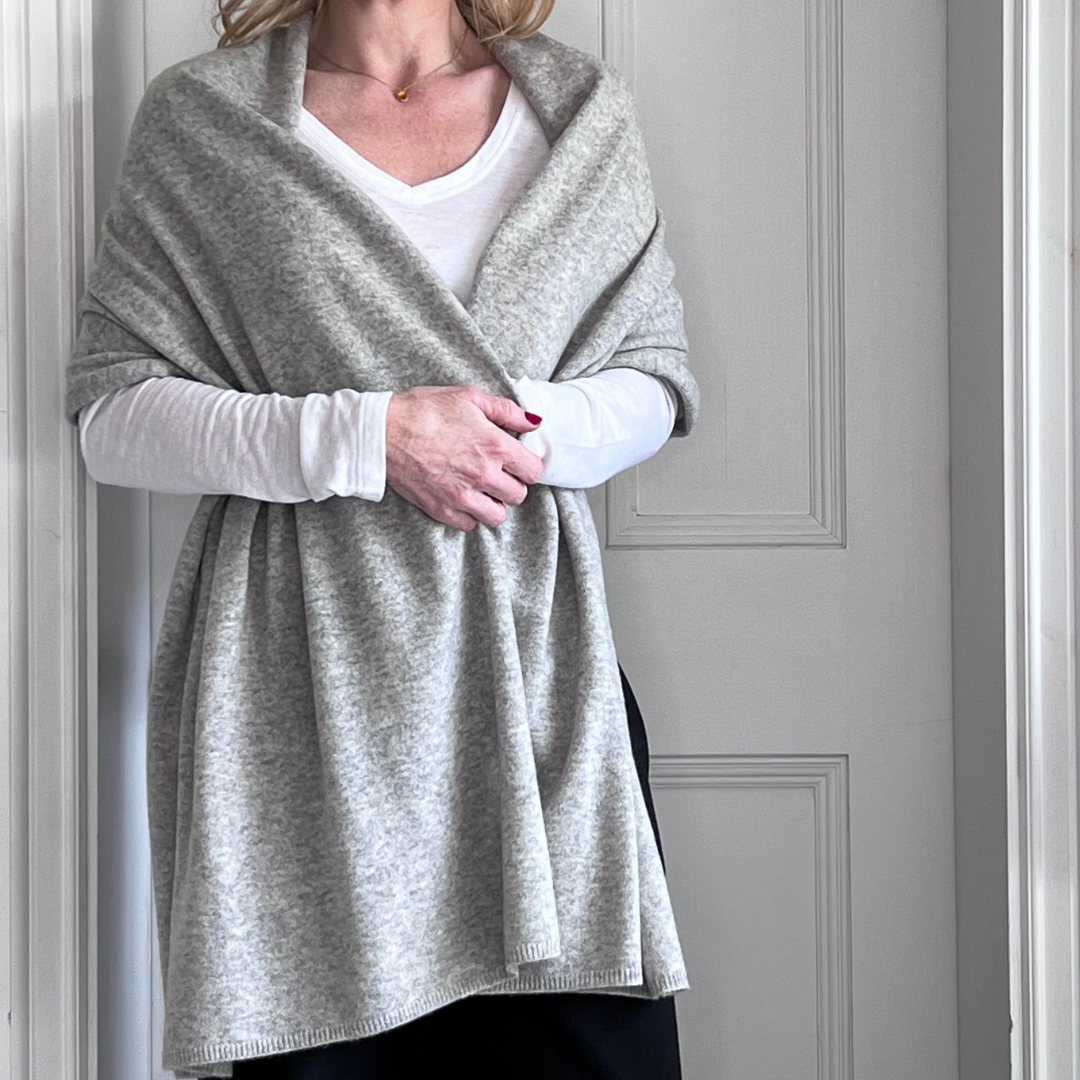 Woman wearing a grey cashmere wrap over a white shirt and black trousers against a white wall.