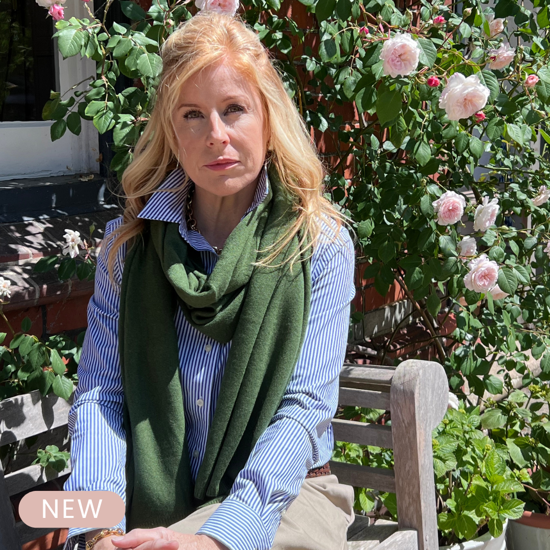 Woman sitting outdoors with green cashmere scarf and floral background. Cashmere travel wrap in forest green.