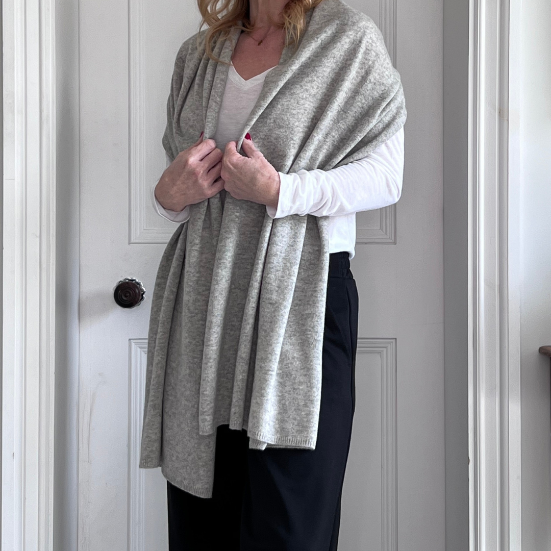 Woman wearing a grey shawl over a white sweater and black trousers in front of a white door.