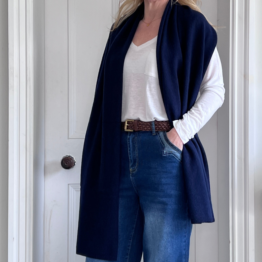 Woman wearing a navy cashmere travel wrap, white shirt and blue jeans standing against a white wall.