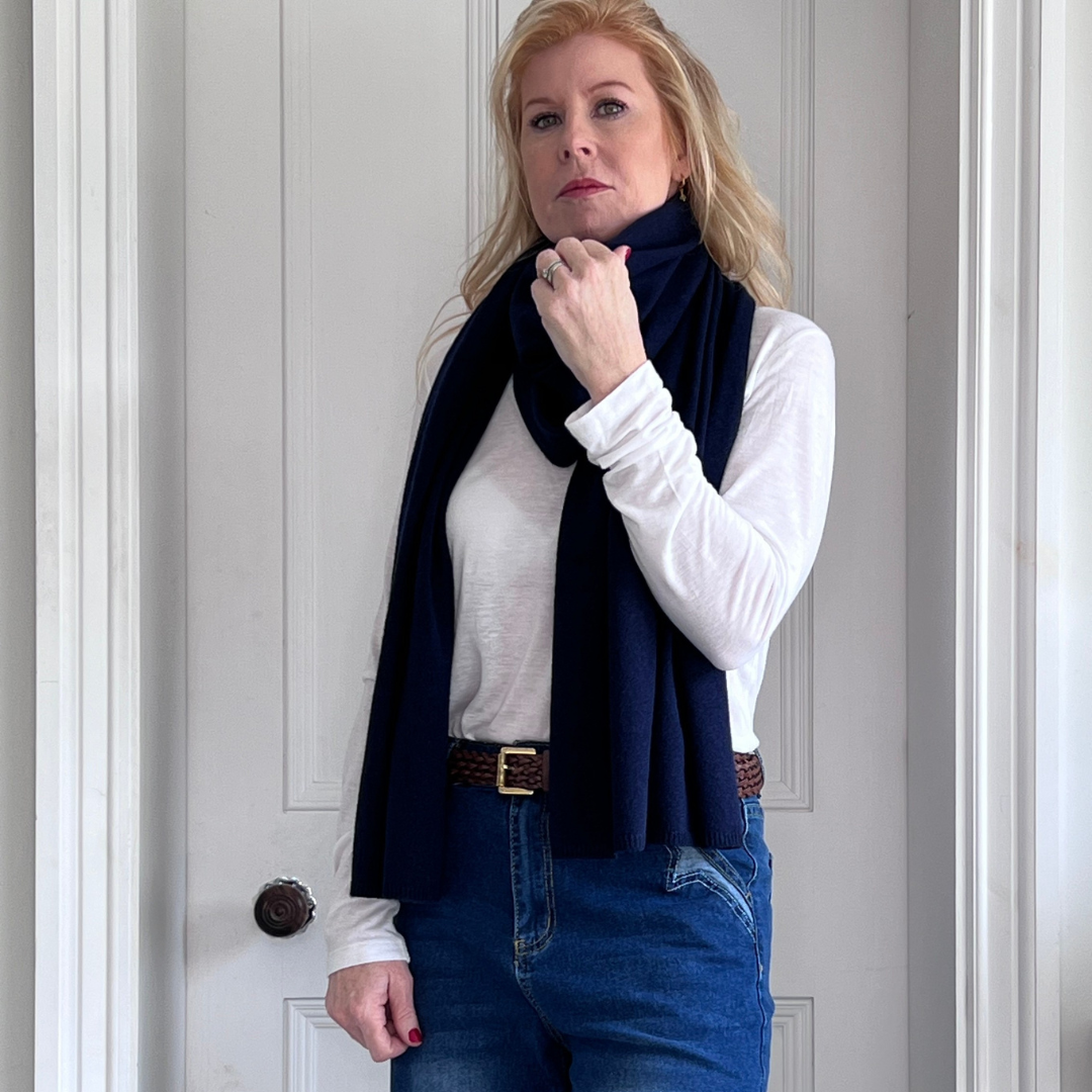 Woman wearing a navy cashmere scarf, white shirt and blue jeans against a gray wall.